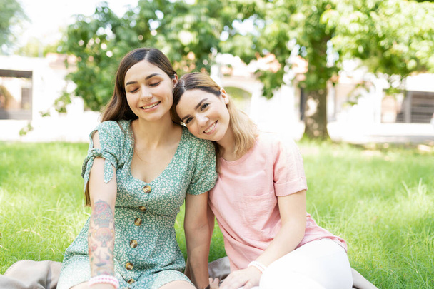 Portret van een lachend mooi LGBT paar met één partner die het hoofd op de schouder van haar vriendin rust en op een deken in het gras zit   - Foto, afbeelding