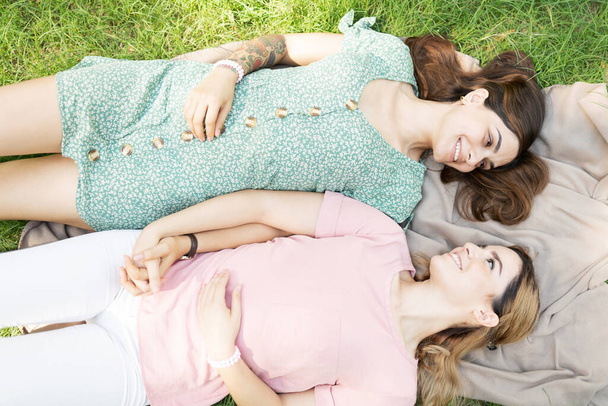 Многорасовая лесбийская пара отдыхает на одеяле в траве рядом друг с другом, держа за руки - Фото, изображение