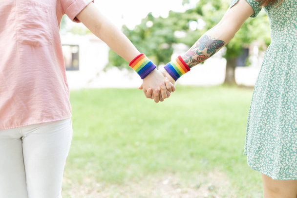 Nahaufnahme von zwei lesbischen Freundinnen, die sich an den Händen halten und Armbänder mit Regenbogenfahnen tragen - Foto, Bild