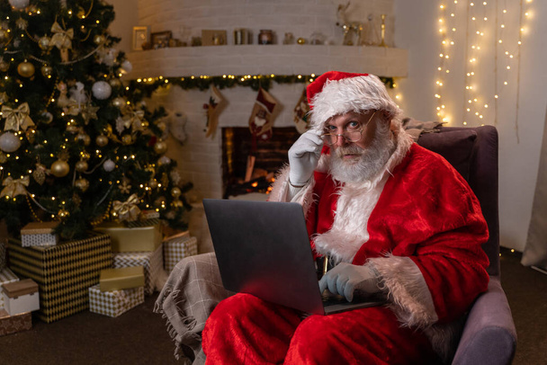 Der moderne Weihnachtsmann. Fröhlicher Weihnachtsmann am Laptop und lächelnd auf seinem Stuhl sitzend mit Kamin und Weihnachtsbaum im Hintergrund. - Foto, Bild