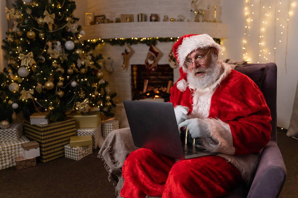Modern Santa Claus. Cheerful Santa Claus working on laptop and smiling while sitting at his chair with fireplace and Christmas Tree in the background. - Foto, imagen