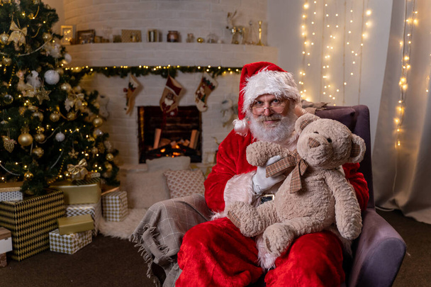 Funny santa claus sitting in his rocker year christmas tree with Teddy bear christmas spirit, holidays and celebrations concept - Foto, imagen