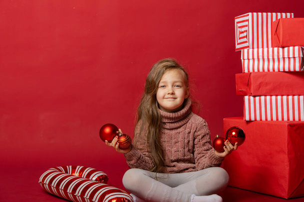 Little cute girl in the studio on a red background with Christmas toys - Фото, зображення