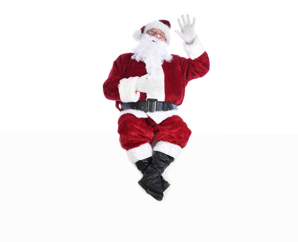 Senior man in traditional Santa Claus Suit sitting on a white wall with one hand in the air and the other on his belly and his eyes closed.  Isolated on white with copy space. - Photo, image