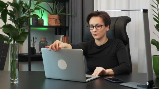 Motivated young businesswoman in glasses is opening laptop and starting to type on keyboard. Focused brunette woman works on computer in her modern office at workplace. Woman is using notebook at work - Video, Çekim