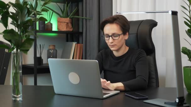 Happy woman in glasses is using laptop computer in modern office. Young businesswoman closes notebook and finishes work at her workplace desk in office, excited with good working project at job - Záběry, video