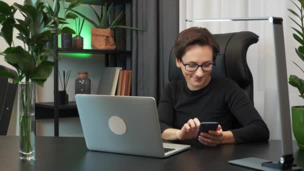 Young smiling woman is using smartphone sitting in modern office. Businesswoman is touching screen of phone, scrolling pages and watching news on internet and social media. Female works at workplace - Séquence, vidéo