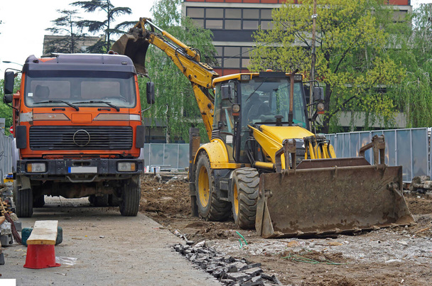 Экскаватор загружает грязь в грузовик на строительной площадке в городе - Фото, изображение