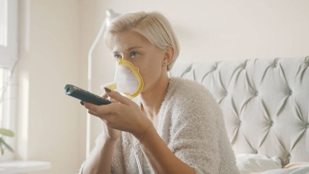 Mujer rubia joven usando inhalador o mascarilla respiradora en su dormitorio. Cambio de canales de televisión - Foto, Imagen