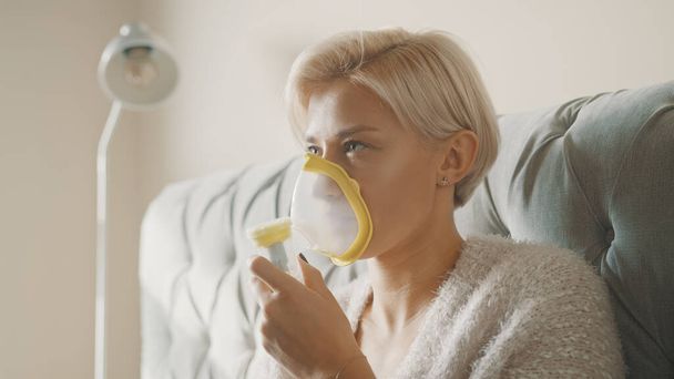 Jonge blonde vrouw met behulp van inhalator of masker in haar slaapkamer - Foto, afbeelding