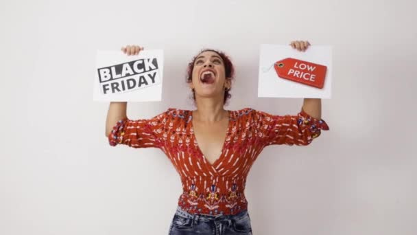 Very happy girl smiling celebrating and holding up two posters of Black Friday and low prices with her hands. Young woman with white skin and curly red hair dressed in colorful patterned clothing - Footage, Video