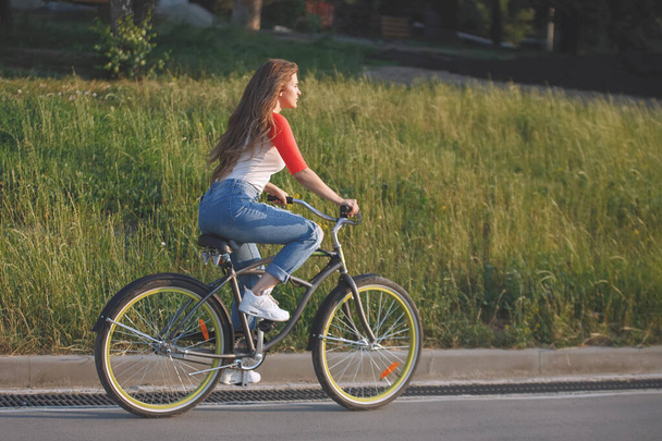 Mulher monta uma bicicleta em uma pista em um parque da cidade. Cabelo comprido, jeans, camisa vermelha e branca. - Foto, Imagem