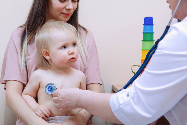 Pediatric lung respiration examination. Baby blonde girl sitting on mom's lap. Female doctor in a medical clinic. - Photo, Image