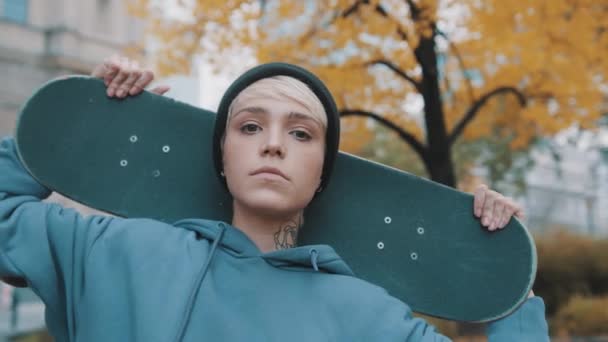 Joven mujer caucásica patinadora sosteniendo monopatín detrás de su cabeza en el parque en otoño - Imágenes, Vídeo
