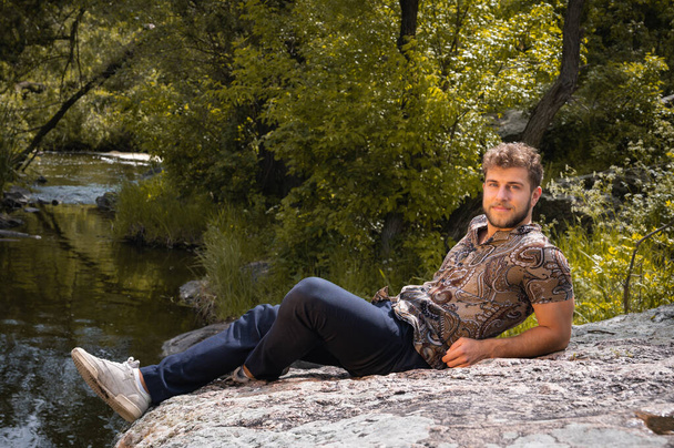 Nuori komea tuhatvuotinen sukupolvi mies on levätä viikonloppuna kalliolla Karst kanjoni lähellä järveä yllään rento vaatteita ja ranta kirjava paita. Vapaa-aika syksyllä, luonnolliset eloisat sävyt - Valokuva, kuva