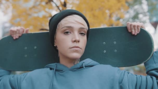 Joven mujer caucásica confiada patinadora sosteniendo monopatín detrás de su cabeza en el parque en otoño - Imágenes, Vídeo