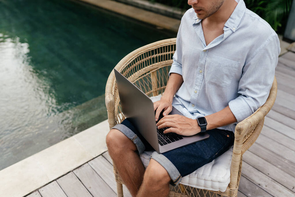 Young man using a laptop computer in a garden with a swimming pool. - Φωτογραφία, εικόνα