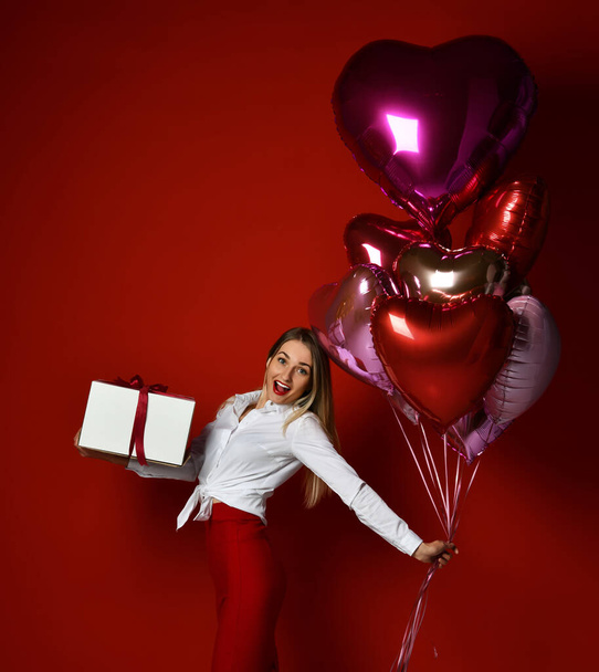 Surprised happy blonde woman in white blouse and red skirt stands with big gift box and colorful balloons - 写真・画像
