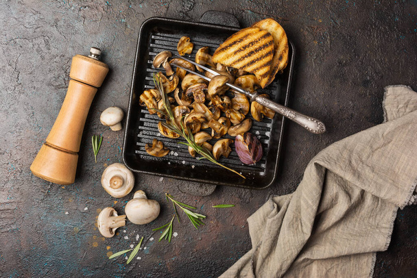 Top view of grilled champignon mushrooms on cast iron skillet or pan with spice, fried onion, rosemary and bread on dark concrete background - Fotó, kép