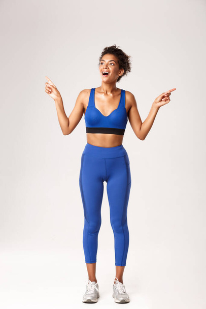 Heyecanlı ve mutlu Afro-Amerikan spor kadın, mavi spor giyer, parmaklarını yana doğru gösterir ama mutlu bir gülümseme ile sola bakar, beyaz arka plan - Fotoğraf, Görsel