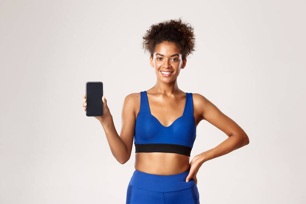 Concepto deportivo y tecnológico. Mujer atlética feliz en ropa de fitness, mostrando la pantalla del teléfono inteligente y sonriendo complacido, de pie sobre fondo blanco - Foto, Imagen