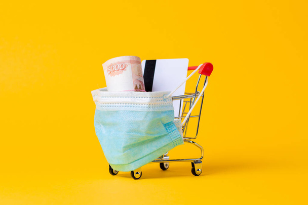 Ein Einkaufswagen mit aufgesetzter medizinischer Maske, gefüllt mit Bargeld und Bankkarte. Gelber Hintergrund. Aus nächster Nähe. Irusschutz und Online-Kauf von Medikamenten. - Foto, Bild