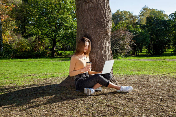 A girl sits near a tree in a park with a cup of coffee and works on a laptop. Freelance work outdoors. Working outside the office due to the Covid 19 pandemic - Fotoğraf, Görsel
