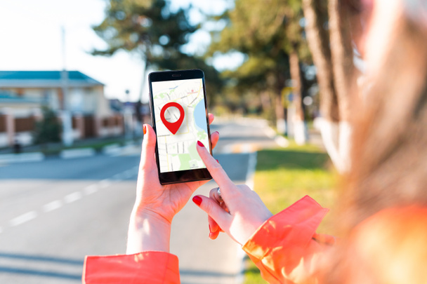 Une main de femme tient un smartphone avec une carte en ligne avec une icône de géolocalisation rouge dessus. En arrière-plan, il y a une rue floue.Gros plan. Concept de navigation en ligne et GPS. - Photo, image