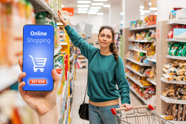 In der Hand hält eine Person ein Mobiltelefon, auf dem Bildschirm liegt ein Einkaufskorb. Im Hintergrund wählt eine Frau verschwommen Lebensmittel in einem Supermarkt aus. Das Konzept des Online-Shoppings. - Foto, Bild