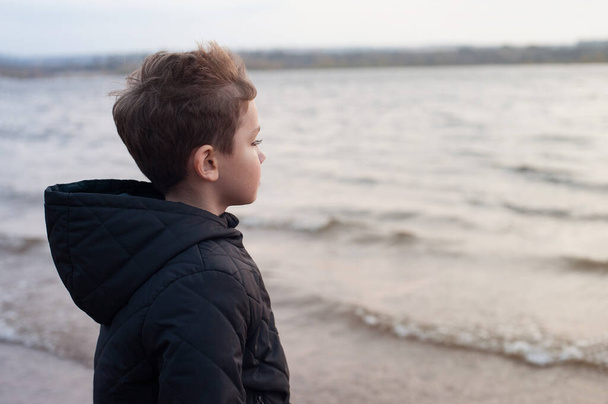 um menino loiro em uma jaqueta escura fica no vento na margem do rio e olha para a distância - Foto, Imagem