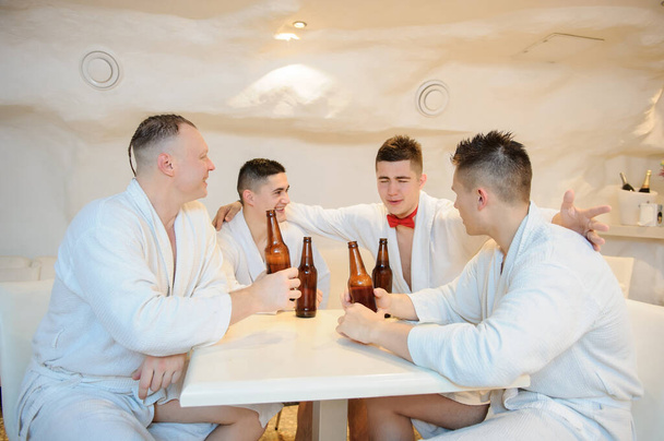 close up photo of 4 men in white gowns sitting around a table and drinking beer after sauna procedures - Photo, Image