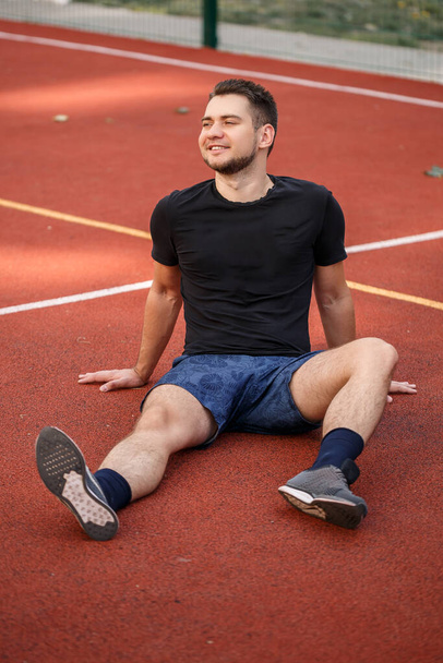 Un jeune athlète masculin barbu est assis sur un court de tennis rouge. Beau mec en t-shirt et short - Photo, image
