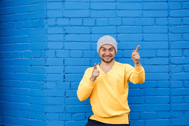Handsome man with a beard and a smile on his face against the background of a blue brick wall. The guy shows emotions to the camera, he is in a yellow sweater. - Photo, Image