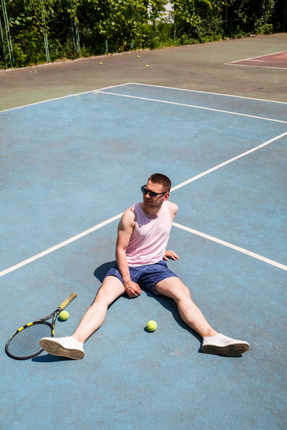 Красивый мужчина в футболке и шортах сидит на теннисном поле с ракеткой. Парень в солнечных очках - Фото, изображение