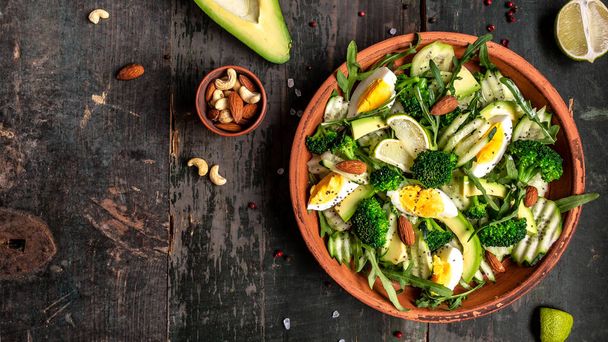 Avocado salade met gekookte eieren, broccoli, komkommer, arugula en amandelnoten in kom op donkere achtergrond. Veganistisch, detox Boeddha schaal, Bovenaanzicht. - Foto, afbeelding