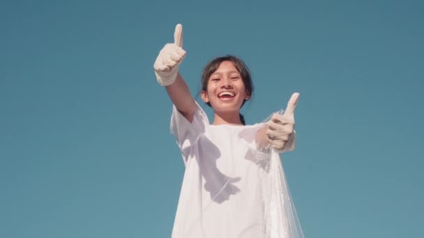 Low-angle footage of teenage indonesian girl wearing white t-shirt and special gloves smiling to camera showing thumbs-up happy to help environment with clear blue sky in background - Кадры, видео