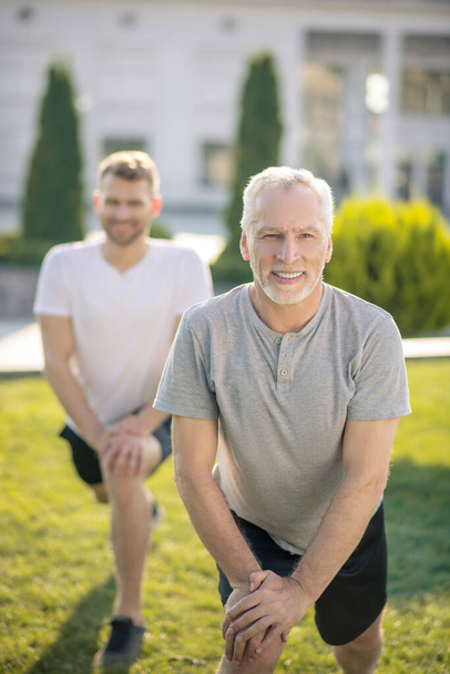 Young male and mature grey-haired male stretching legs outside, hands on knee, smiling - Foto, Bild