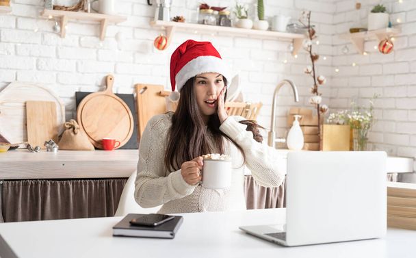 Mladá překvapená brunetka žena v Santa klobouk chatování s přáteli pomocí svého notebooku v kuchyni - Fotografie, Obrázek