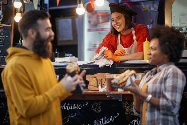 caucasian female employee in fast food service smiling, looking at beardy hipster talking with a male friend - Fotoğraf, Görsel