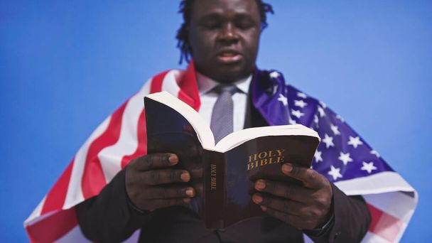 Κοντινό πλάνο, Αγία Γραφή στα χέρια του μαύρου αφροαμερικανού άνδρα με κομψό κοστούμι με σημαία των ΗΠΑ πάνω από τους ώμους - Φωτογραφία, εικόνα