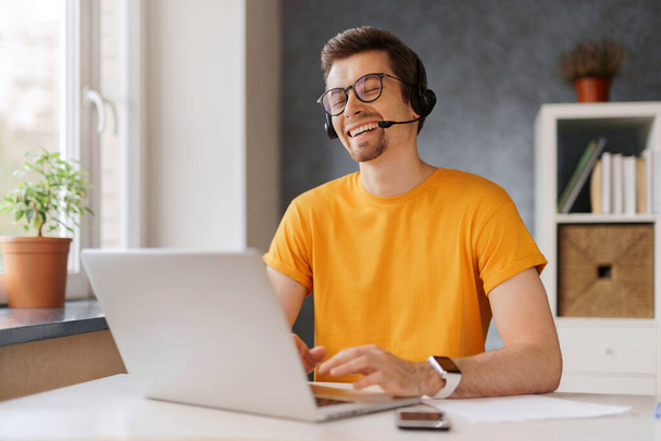 ヘッドセットを着用した幸せな笑い男は友人や家族との会議ビデオ通話で通信します。自宅からノートパソコンで遠隔作業をしているフリーランサーは笑顔。オンラインクラスのウェビナーコースがあります - 写真・画像