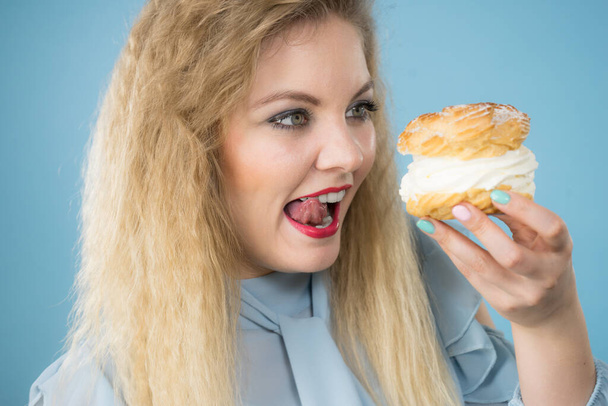 Sweet food and happiness concept. Funny joyful blonde woman holding yummy choux puff cake with whipped cream, excited face expression. On blue - Photo, Image
