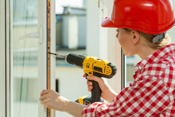 Young woman wearing helmet using power drill and tools for work at home, installing windows. Girl working at flat remodeling. Building, repair and renovation. - 写真・画像