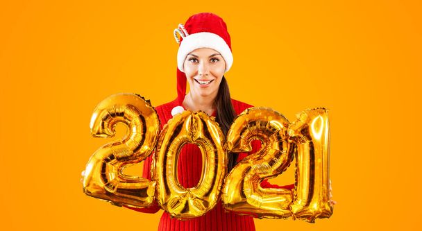 New Year 2021 Gold Balloons. Young woman in Santa Claus hat having smiling holding in hand 2021. Girl in red hat and red Dress stands on bright orange background. Concept Christmas celebration - Zdjęcie, obraz
