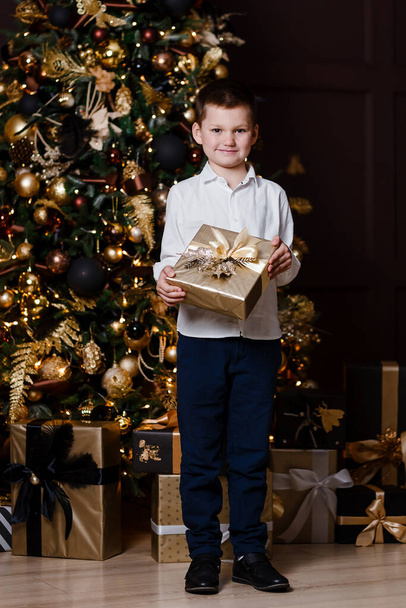 A boy in a white shirt and trousers holds a Christmas present in his hands near the Christmas tree in full height - 写真・画像