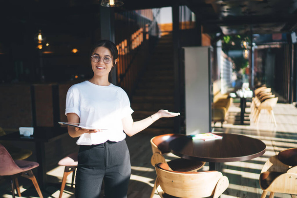 Jeune femme gestionnaire positive dans les lunettes et t-shirt blanc souriant tout en démontrant l'intérieur de la cafétéria moderne avec tablette à la main - Photo, image