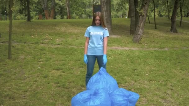 Portrait of tired beautiful volunteer wiping forehead with hand in protective gloves. Wide shot of exhausted slim Caucasian woman posing in clear summer park with garbage bags. Eco-protection. - Metraje, vídeo