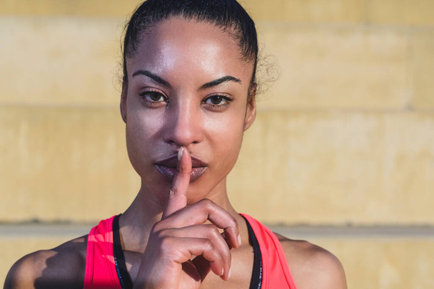 portrait of attractive young African woman wearing sportswear looking at camera and making silence gesture with a finger on her lips. copy space on the right - Photo, Image