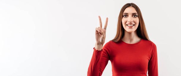Портрет счастливой молодой женщины с длинными каштановыми волосами с жестом мира, носящей обычный красный свитер. Студийный снимок, белый фон. Человеческие эмоции, концепция выражения лица - Фото, изображение