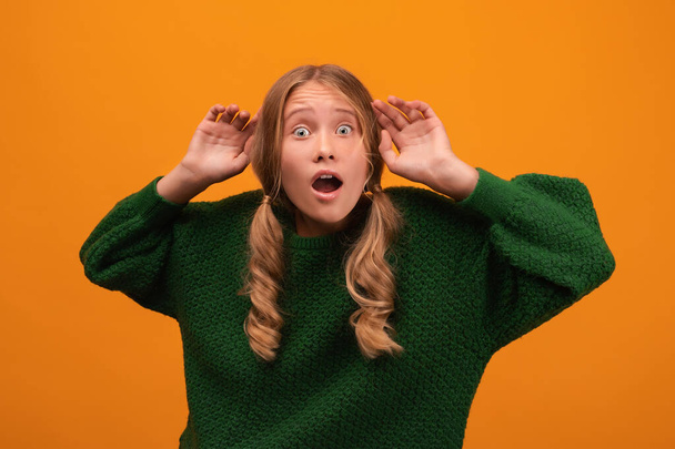 Image of shocked blonde girl 12-14 years old in warm green sweater expressing surprise on camera. Studio shot, yellow background, isolated. Human emotions, facial expression concept - Photo, Image